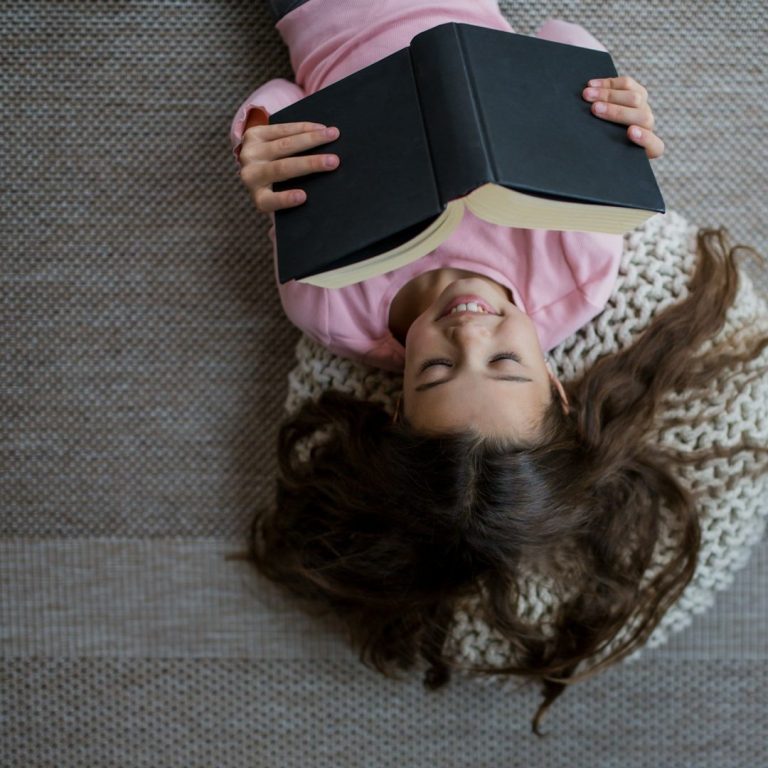 Happy reading A girl in a pink sweater lies on a cozy rug, smiling while reading a book.