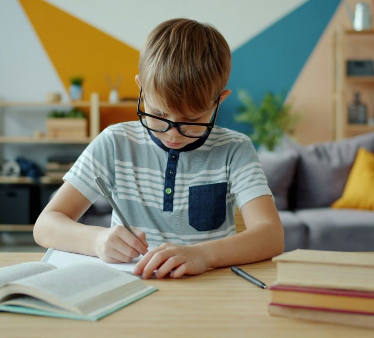Boy studying A boy wearing glasses writes in a notebook at a desk in a brightly decorated room.